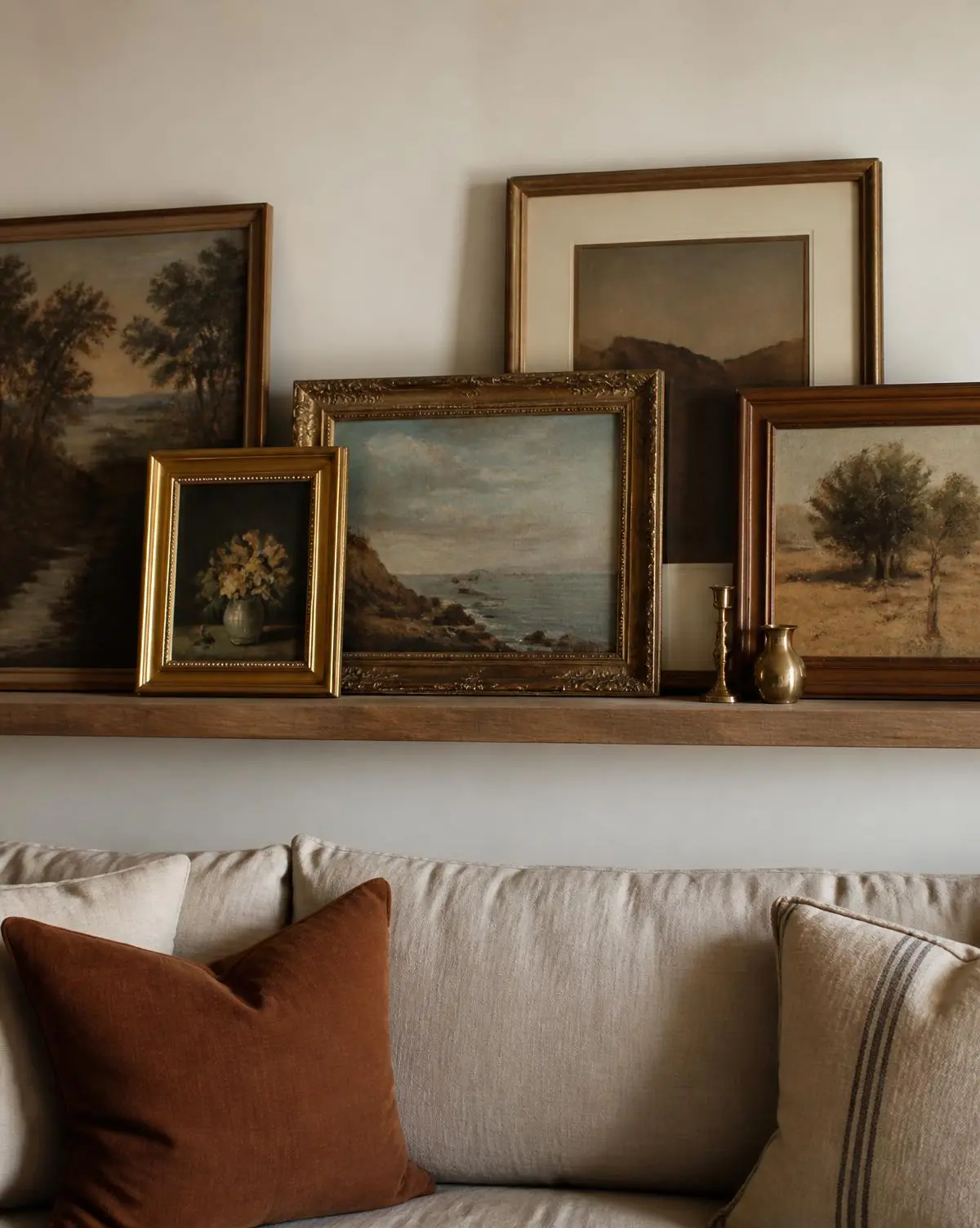 Vintage paintings layered on a wooden picture ledge above a linen sofa